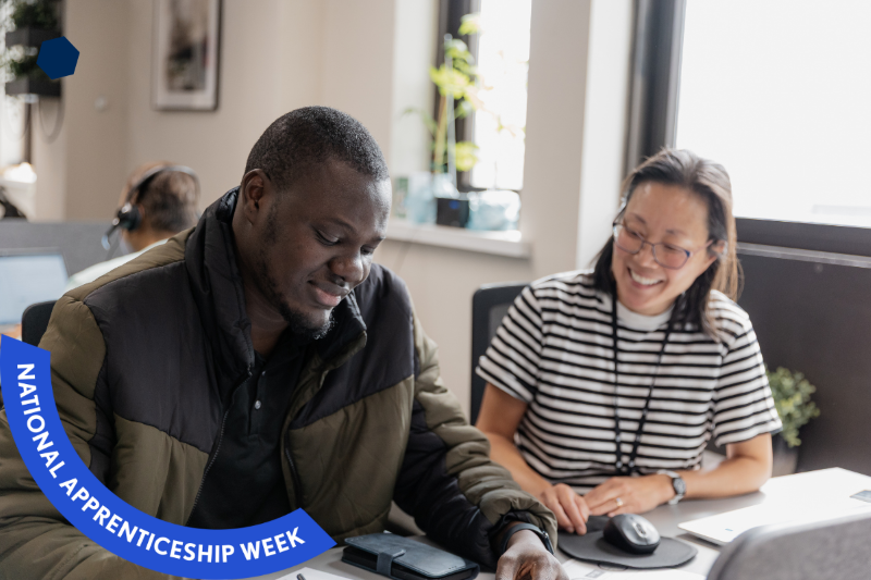 Two colleagues working collaboratively and smiling. In the bottom left there is a blue banner with white text that reads: National Apprenticeship Week