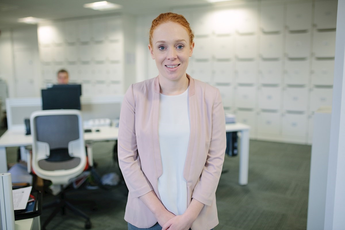 Clare Fallon wearing a pink blazer and white top standing in our office with computers and desks visible in the background