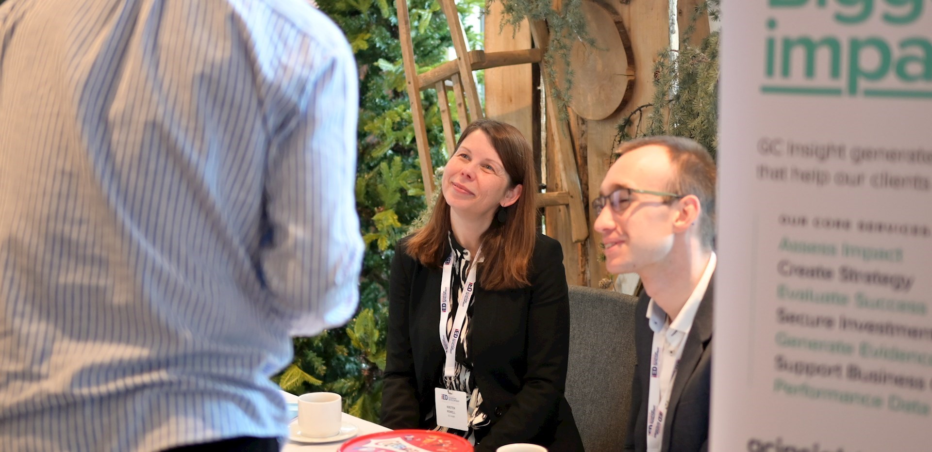 Two GC Insight colleagues at an event sitting next to a GC Insight banner and looking up at an attendee who is speaking to them